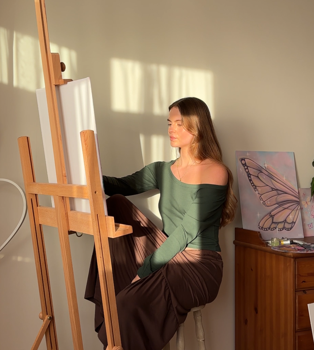 Woman painting at an easel in a home studio with a painting and desk in the background.