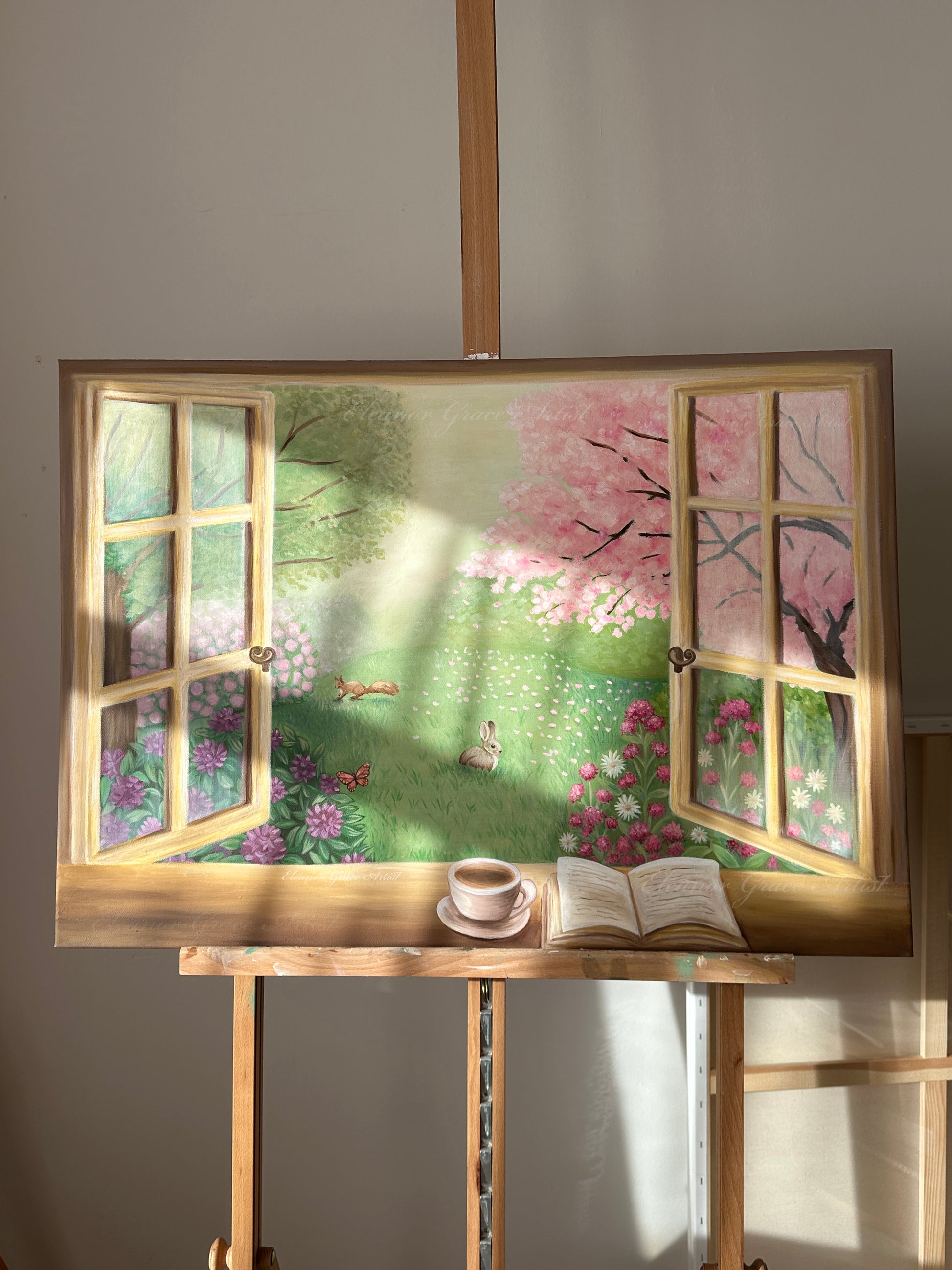 Painting of a garden scene with a window frame, cup, and book on an easel.